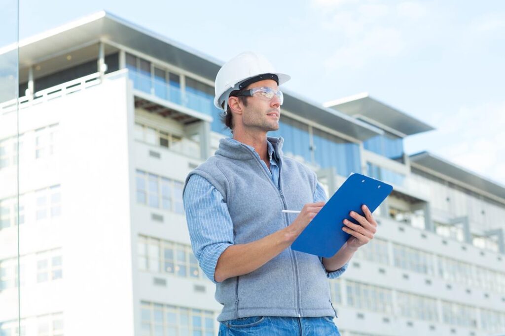 Male architect looking away while writing on clipboard against building
